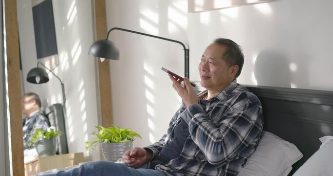 Mature Asian man propping back, raising smartphone and recording voice message on bed to send