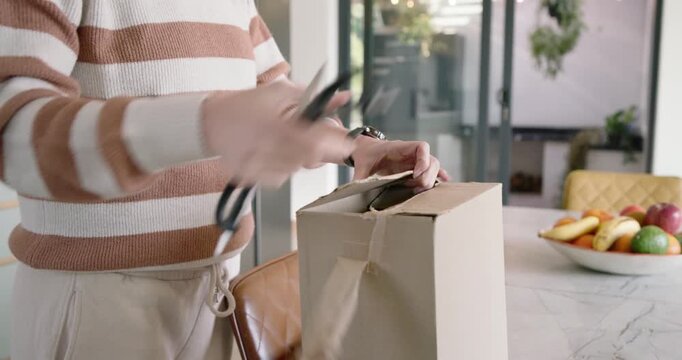 Adult woman unpacking delivery in kitchen, cutting tape with scissors and cutter, inspecting bottle