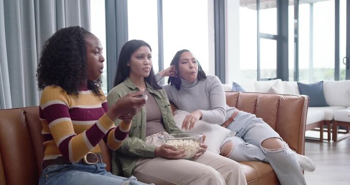 African American women reaching for popcorn, passing remote, leaning together on couch watching TV
