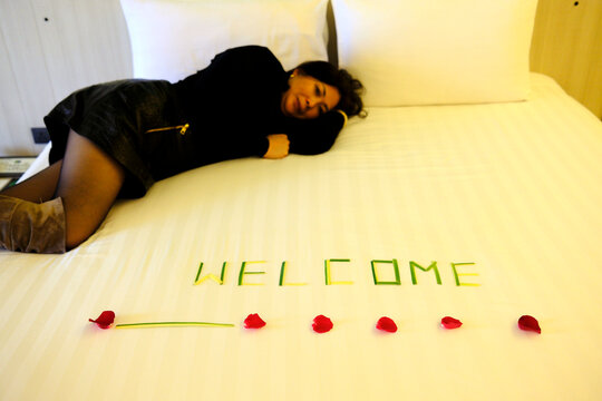 A woman relaxes on a hotel bed where the word "Welcome" is spelled out in rose petals. Pattaya, Thailand.