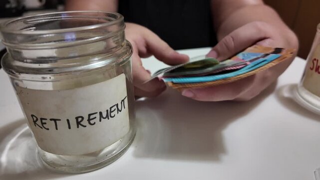 Qawra, Malta 05.10.2026 - Hands hold cash near a labeled jar. Jar marked "RETIREMENT" sits on a table. Fingers gently manage folded bills. Person shows financial preparedness for future. Ideal for ret