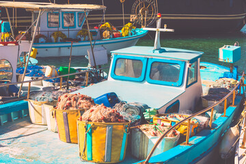 Fishing boats moored in a harbor with nets piled in crates and baskets on the deck of a small vessel. Wheelhouse has framed windows, and ropes and equipment are scattered along the railings © Leilani
