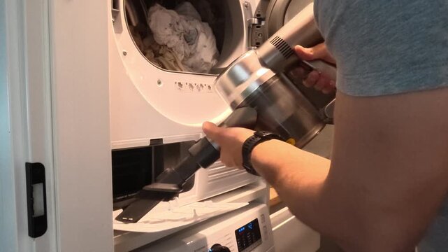 Man performing routine maintenance of a household tumble dryer, removing lint and dust from the filter compartment using a vacuum cleaner, cleaning brush and cloth. Domestic appliance cleaning and hom