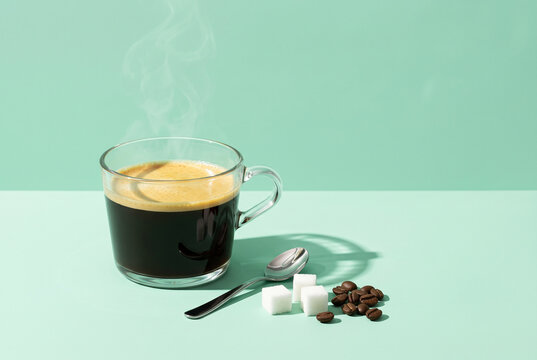Steaming hot black coffee in a clear glass cup with sugar cubes and beans on a minimalist mint green background.