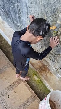 A young boy wearing a dark long-sleeve swimwear leans forward under an outdoor water tap, washing his head and hair after swimming.