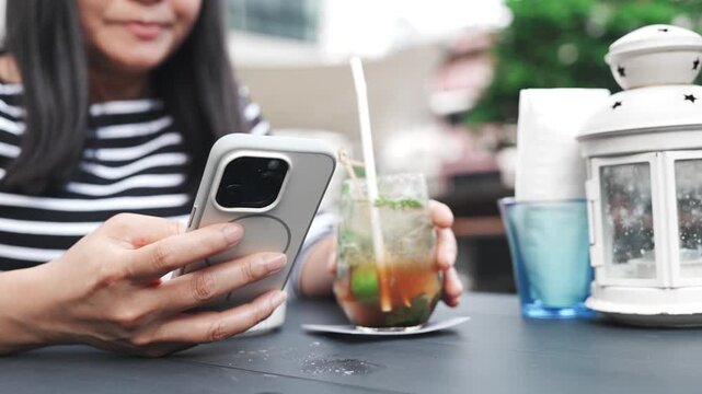 Woman using smartphone at outdoor cafe, talking while typing, sipping iced drink and eating snacks, middle aged female enjoying casual conversation, leisure time and relaxing lifestyle during break.