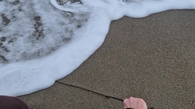 Person drawing shape in wet beach sand with twig while ocean waves wash ashore nearby. First-person perspective point of view shot.