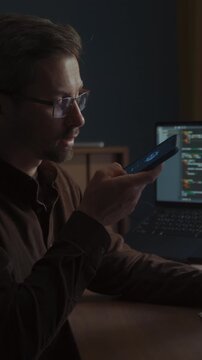 A programmer working from home office using an AI voice assistant app on his smartphone to multitask, while an artificial intelligence agent generates program code on his computer. Vertical video