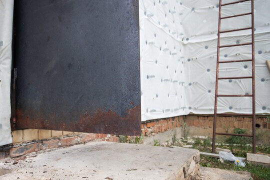 rusted metal entrance door left open beside an unfinished insulated facade and ladder, showing aging housing, deferred maintenance and residential renovation needs