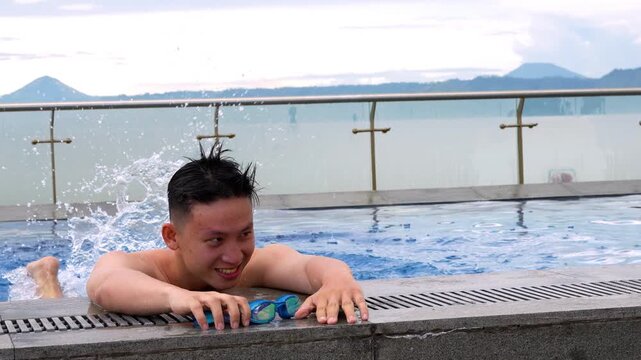 happy young Asian man relaxing at the edge of a swimming pool holding goggles, with a scenic mountain background