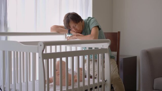 Sleepy father stroking five-month-old son in crib while falling asleep. Tender family moment.