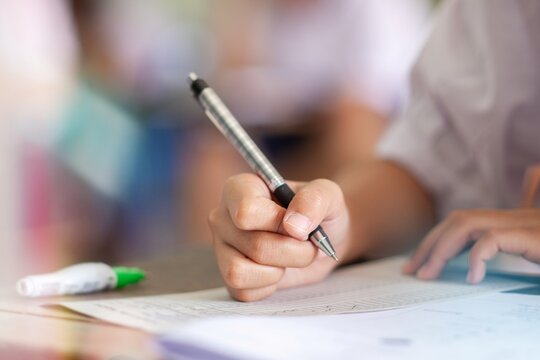 Close-up of a student's hand diligently writing on an exam paper during a school test. 
The student is using a professional pen , sitting at a wooden desk in a brightly lit classroom.