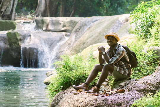 Happy African Male Traveler Sitting and Relaxing Near Waterfall in Nature Park