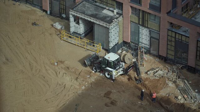 Heavy machinery excavating a deep pit on a construction site, while a worker in overalls stands by and supervises the earthmoving operation during a sunny day. Powerful excavator bucket extracts groun