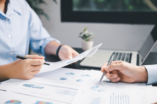 Two coworkers review printed charts with pens beside a laptop at an office desk, discussing business data, report findings, performance trends, and collaborative planning in a bright workspace.