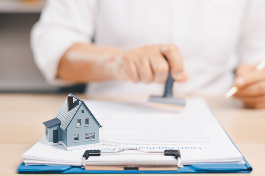 Miniature house on a loan application form as a person uses a stamp symbolizes mortgage approval, property financing, credit review, lending decisions, home purchase, documentation,