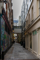 Narrow cobblestone alleyway between tall buildings with an overhead bridge in a city. © DriveAndDive
