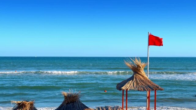 Red warning flag and straw umbrellas on sandy beach. Coastal landscape shows sea waves crashing behind traditional sunshades under cloudless sky.