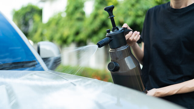 Close-up of woman's hands spraying water from foam pressure sprayer on car hood outdoors. DIY car wash, auto detailing and vehicle maintenance concept.