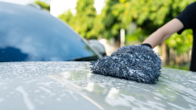 Close-up of person using microfiber wash mitt to clean soapy car hood outdoors. DIY car wash, auto detailing and vehicle maintenance lifestyle concept.