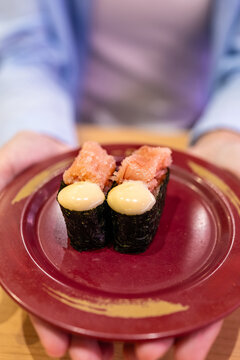 Close-up of woman's hands holding plate with spicy mentaiko mayo gunkan sushi in Japanese restaurant. Delicious gourmet food concept. Vertical Shot