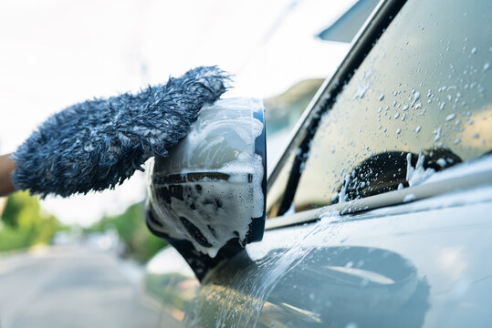 Close-up of hand using microfiber wash mitt to clean soapy car side mirror outdoors. DIY car washing, auto detailing and vehicle maintenance lifestyle concept.