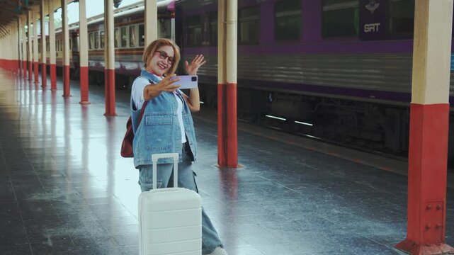 A cheerful mature woman traveler in denim stands on a train platform with her white suitcase and takes a selfie while waving joyfully at the camera as a purple train waits behind her capturing the exc