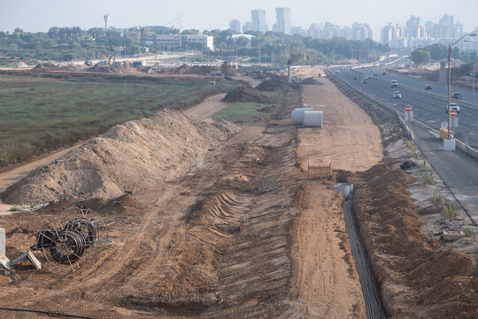 construction zone featuring ground excavation, laid utility cables, and earthworks alongside a multi-lane highway near Netanya, Israel. Modern high-rise buildings and urban infrastructure