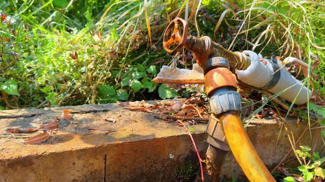 Rusty metal water tap with hose in garden. Old rusty water tap with connected orange hose dripping water onto concrete block.