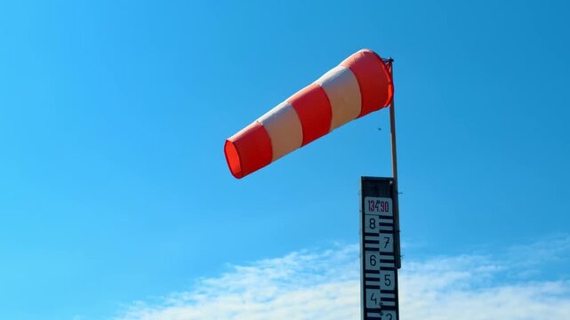 Red and white windsock blowing in wind. Striped red and white windsock mounted on pole with water level gauge against blue sky.