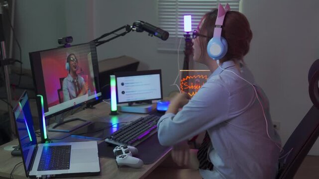 Young woman gamer livestreaming online at her neon lit home studio wearing cat ear headphones while chatting with viewers and reacting to live comments on screen during a late night session