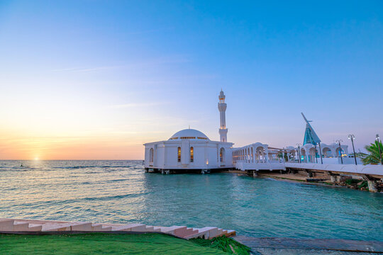 Al Rahmah Mosque, a striking white building with a tall minaret, standing on Jeddah Corniche and appearing to float above the Red Sea as sunset casts vibrant colors across the sky in Saudi Arabia