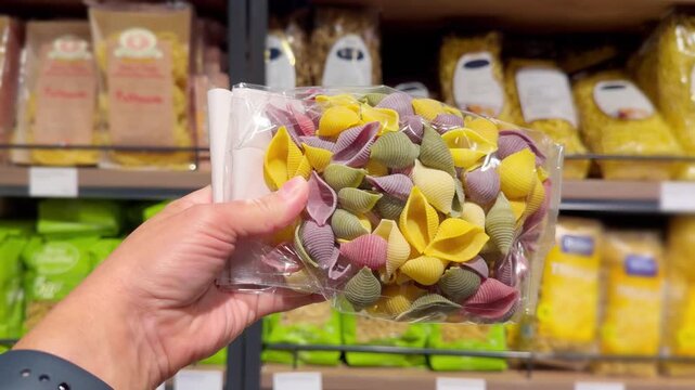 Hand Holding Pack of Colorful Pasta in Supermarket &ndash; Assorted Colored Macaroni Against Blurred Shelves Background