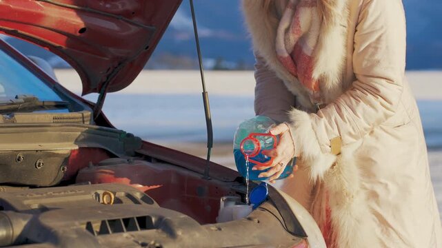 Woman pouring anti freeze windshield fluid into car on cold winter day, snow covered fields and mountains blurred background, closeup to hands holding bottle with blue liquid