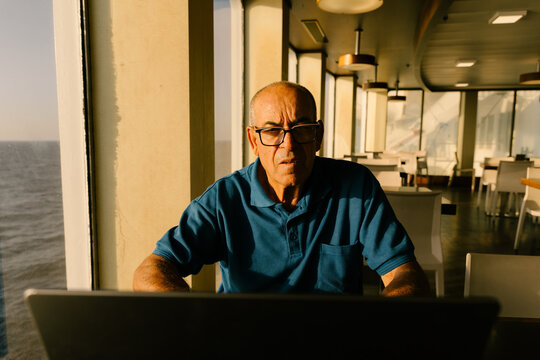 Elderly man taking a video call by a ship window in sunset light.
