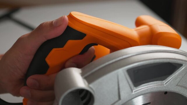 Close-up of hand operating orange and black circular saw handle showing finger on trigger and metal blade guard in workshop lighted setting with textured rubber grip for control and safety purposes.