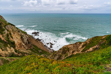Fototapeta premium Cabo da Roca dramatic coastal cliff overlooking Atlantic Ocean in Portugal. Scenic westernmost point of Europe with rugged landscape, ocean waves and natural coastline.
