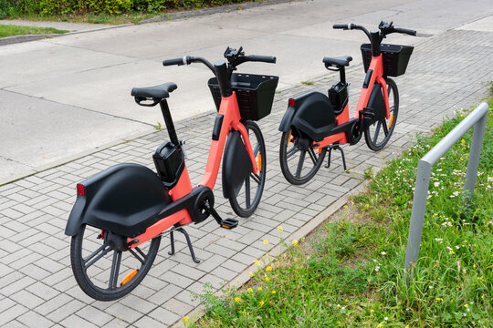 Red shared electric bikes parked beside a bench near a playground in a city park. Urban mobility, shared transport, sustainability, commuting, family leisure, outdoor recreation.
