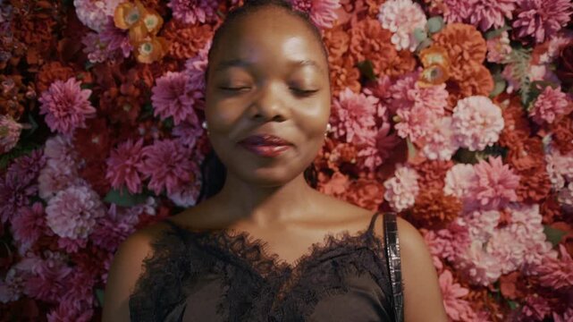 A young Black woman looks directly into the camera with a shy and embarrassed expression. A colorful flower wall creates a vibrant backdrop for this candid portrait.