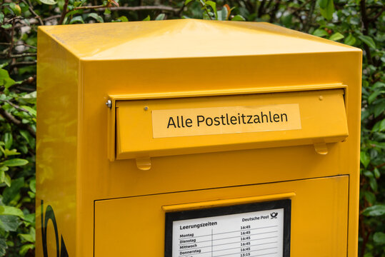 Berlin, Germany - 16 August 2026. A yellow German mailbox collects letters outdoors next to green bushes. Public postal service, mail delivery, communication, community, infrastructure.