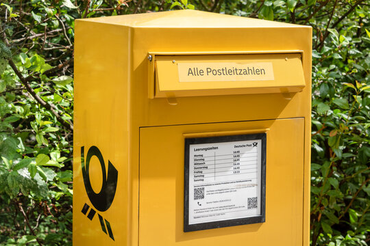 Berlin, Germany - 16 August 2026. A yellow German mailbox collects letters outdoors next to green bushes. Public postal service, mail delivery, communication, community, infrastructure.