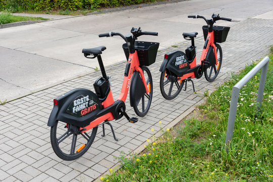 Berlin, Germany - 16 August 2026. Two orange electric rental bikes parked on a city sidewalk next to a grassy verge. Urban bike sharing, electric mobility, sustainable transport, city lifestyle.
