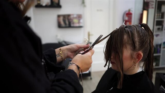 Young female client having her brown hair cut by a professional hairdresser in a modern beauty salon. The stylist carefully trims a strand of wet hair using scissors and a comb for a precise result