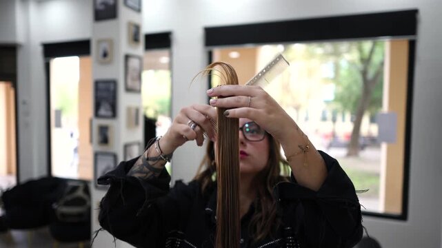 Talented female hairstylist with tattoos carefully cutting a client's long brown hair with professional scissors and a comb in a modern beauty salon, creating a new stylish haircut