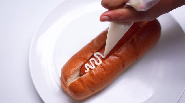 Hand Drizzling White Sauce in Zigzag Pattern Over Hot Dog Bun During Food Preparation.