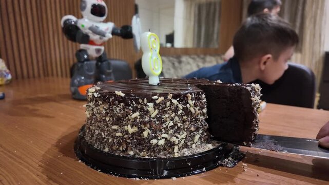 Child cuts birthday cake with knife. Robot toy stands nearby on table. Child watches with interest. Chocolate cake covered in chopped nuts. Warm, joyful family celebration moment