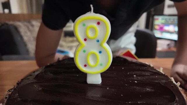 Child blows out birthday candle shaped like number eight. Chocolate cake with nuts sits on wooden table. Child smiles, happy, during celebration. Perfect for birthday, child, celebration, family