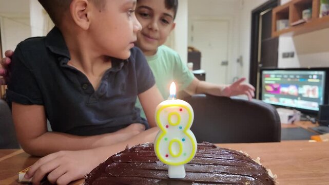 Happy birthday boy blows out candle on chocolate cake. Young child smiles as he blows out birthday candle. Another child watches with joy in background