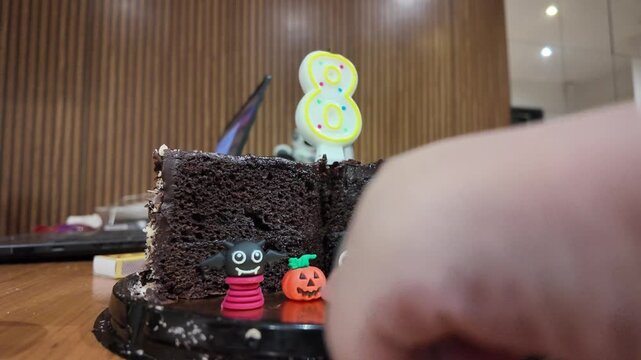 Child's hand reaches for chocolate cake with Halloween decorations. Candle shaped like number eight glows softly. Small pumpkin and skull figures sit beside cake. Cake slice appears festive and sweet