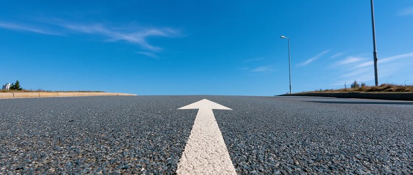 White directional arrow painted on textured asphalt road surface under bright clear blue sky with scattered wispy clouds, solitary streetlight pole